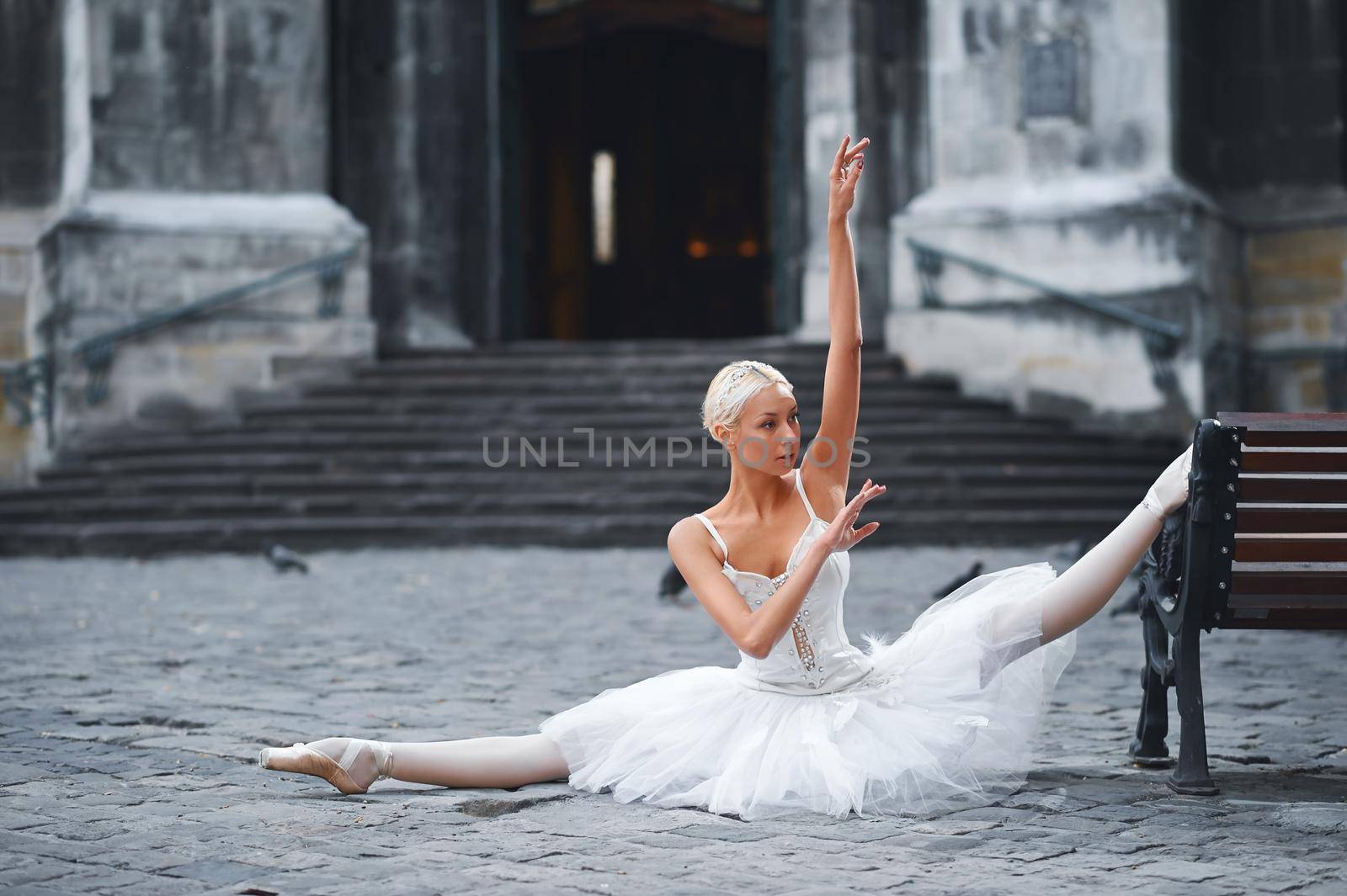 Horizontal shot of a beautiful ballerina performing in the city center outdoors doing splits near the bench stretching flexibility balance beauty athletics urban fashion grace concept.