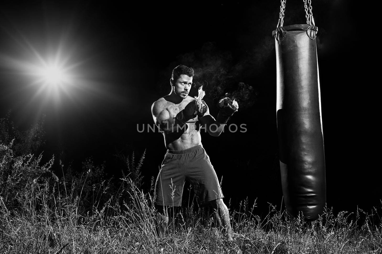 Monochrome portrait of a focused male boxer exercising outdoors hitting a punching bag wearing burning boxing gloves copyspace concentration determination fit toned body torso strengthening concept.