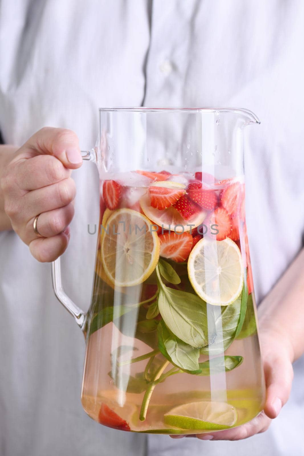 A jug with a refreshing organic soft drink with ripe fresh strawberries, lemon slices with lime and basil, in female hands close-up.