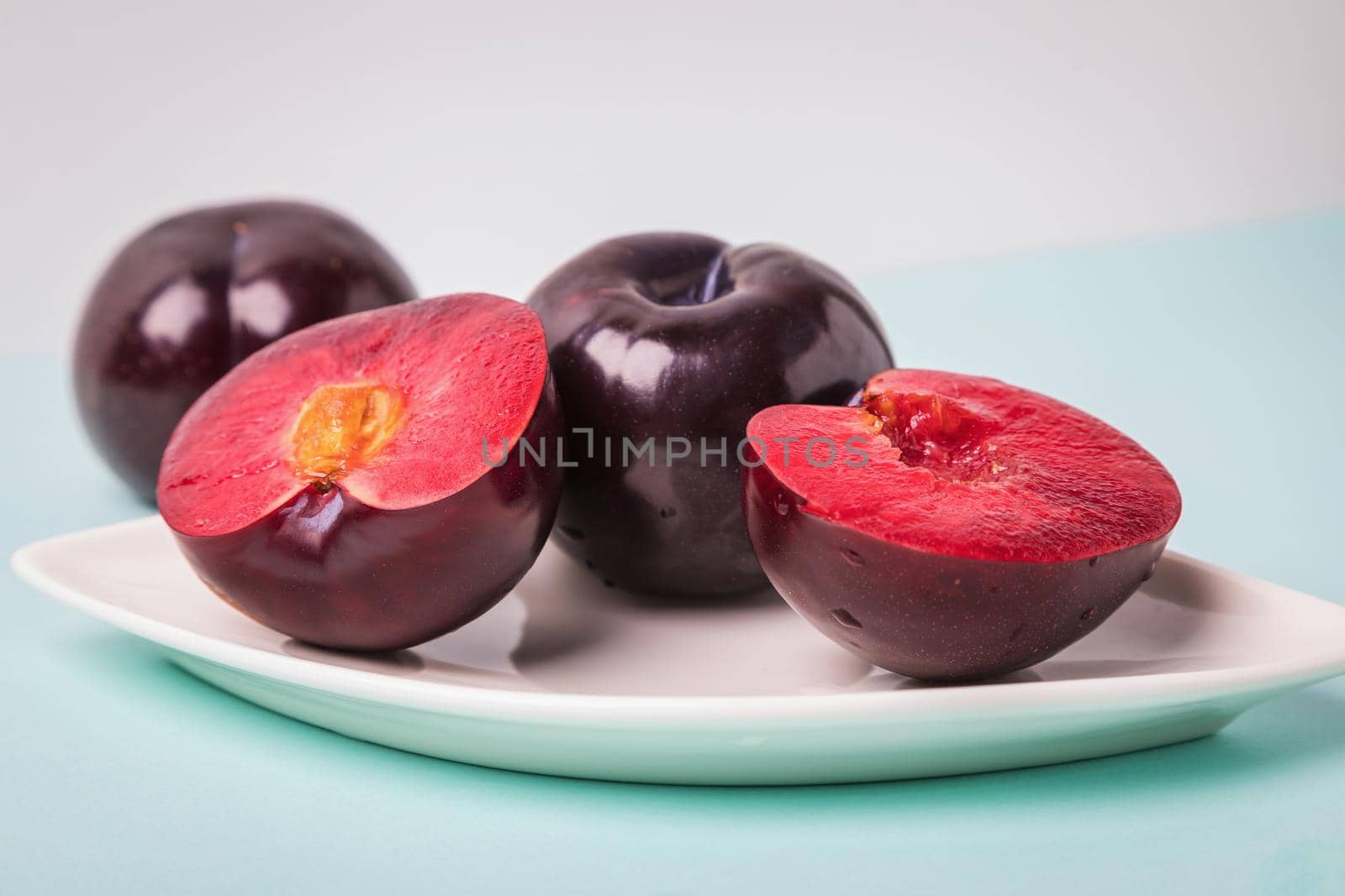 On a white saucer is a large plum, cut in half, and next to it is a whole plum. The pulp of red berries. Close-up.