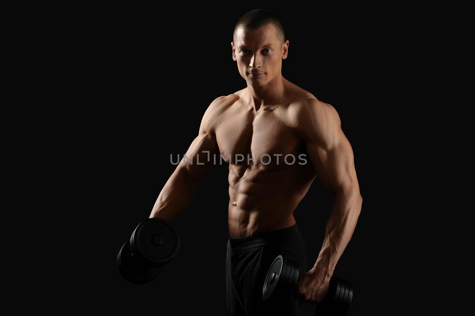 In the name of perfect body. Horizontal studio shot of a young bodybuilder man posing with dumbbells smiling to the camera posing shirtless with his fit and toned torso