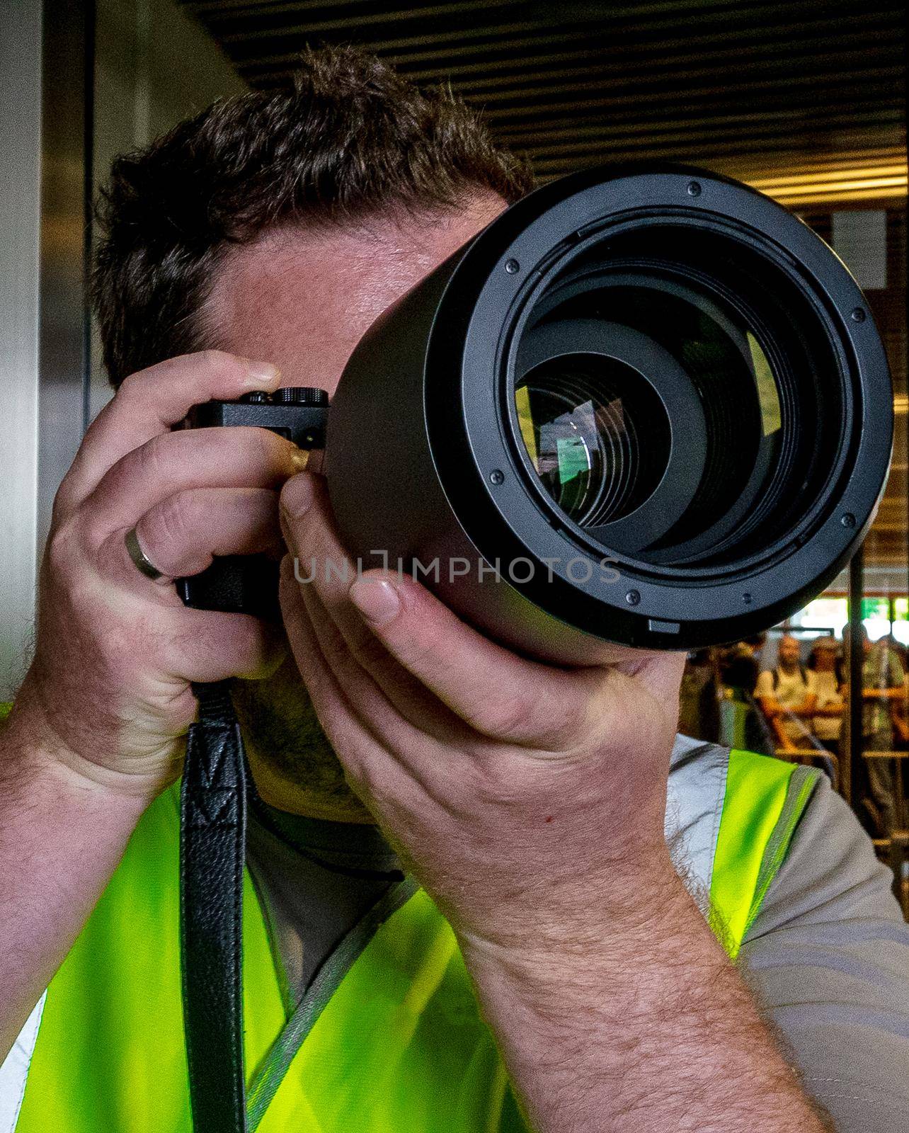 A male photographer in a green reflective vest looks through the camera's viewfinder.