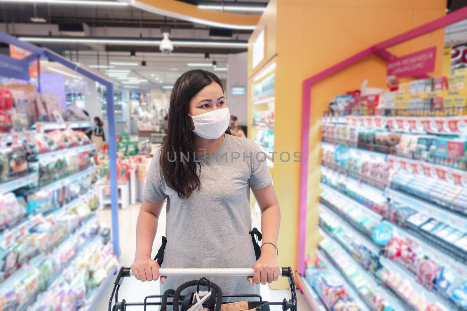 Customer Asian Woman Wearing Face Mask With Shopping Cart in Supermarket Department Store Shop While Choosing and Looking Something on Shelf During Covid-19 Pandemic. Coronavirus Covid Situation