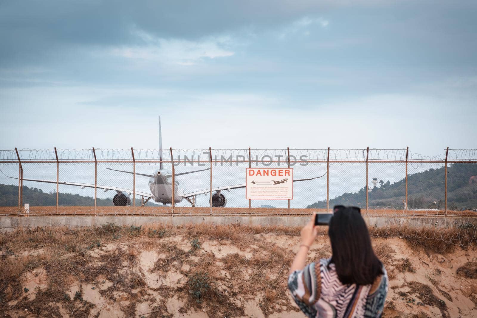Tourist Woman is Capturing Photos The Airplane While Take off Landing on Runway Track From Outside The Airport. Asian Tourist Woman Having Fun With Photographic Vehicle Aircraft Beside Airport Fence. by MahaHeang245789