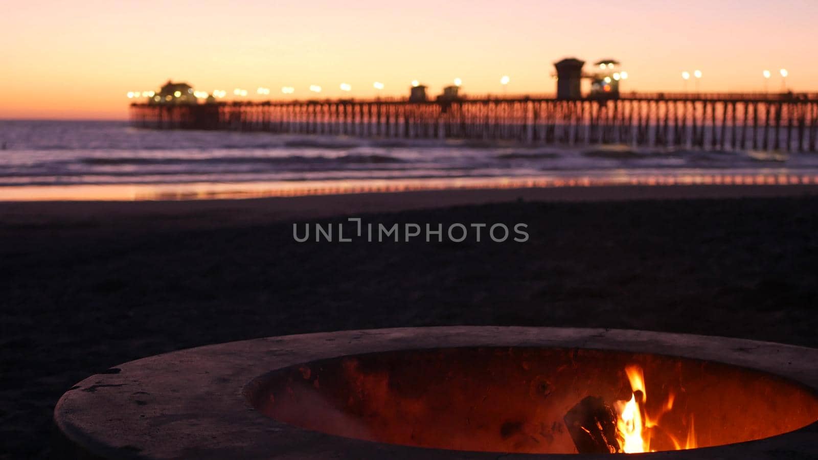 Campfire pit by Oceanside pier, California USA. Camp fire burning on ocean beach, bonfire flame in cement ring place for bbq, sea water waves. Romantic evening twilight sky, dusk after summer sunset.