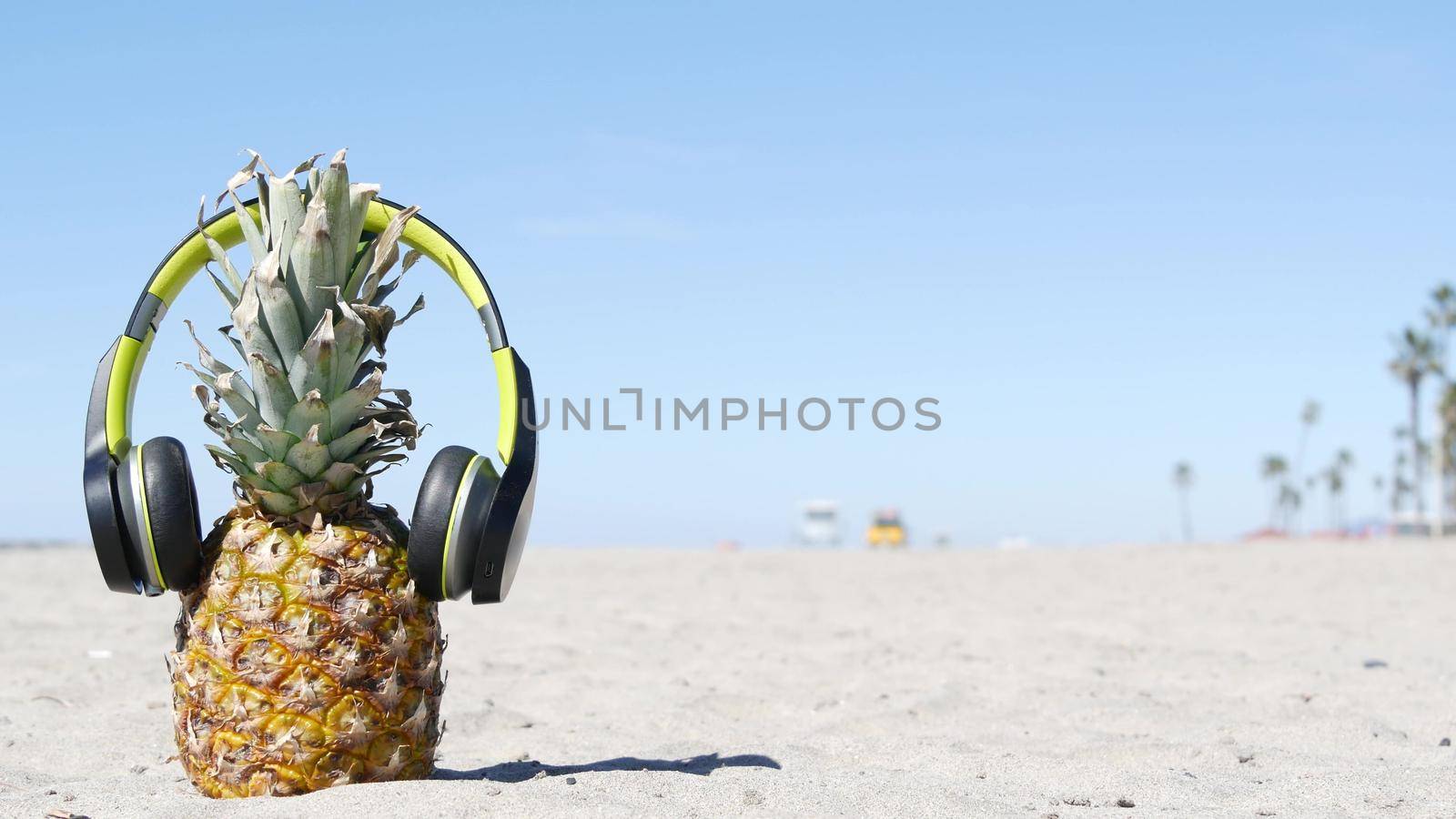 Funny pineapple in headphones, sandy ocean beach, yellow lifeguard car, California USA, Pacific coast. Tropical summer exotic fruit enjoying vacations and music in paradise. Ananas sunbathing on shore