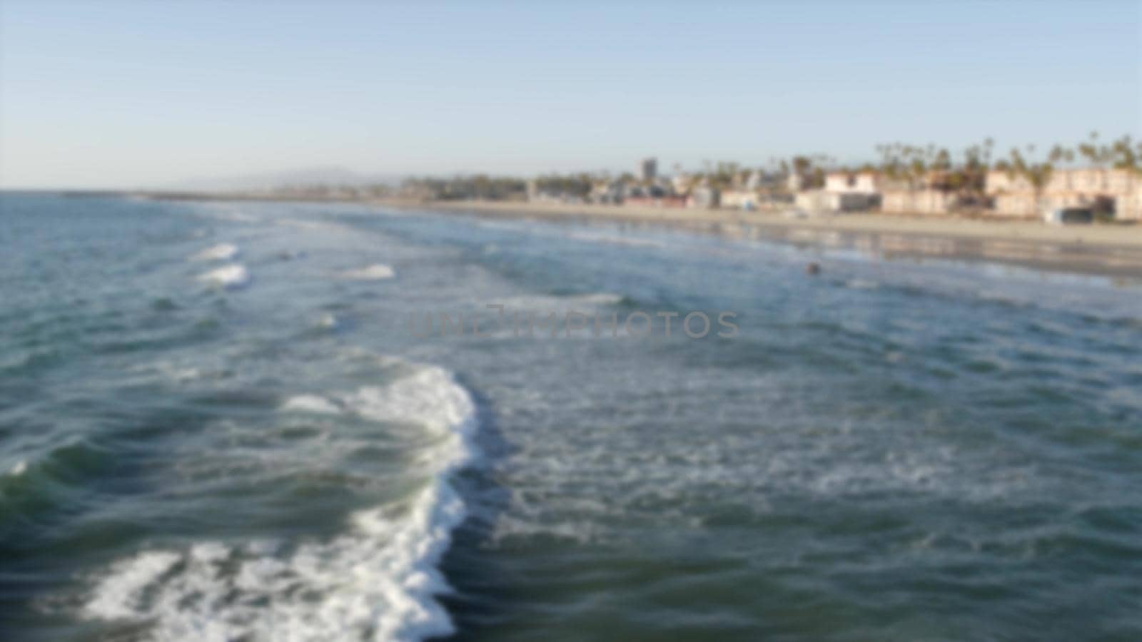 Defocused pacific ocean coast from pier. Sea water waves tide, shore sand. Beachfront vacations resort. Waterfront Oceanside, California USA. Tropical palms and houses on beach. Reflection in littoral