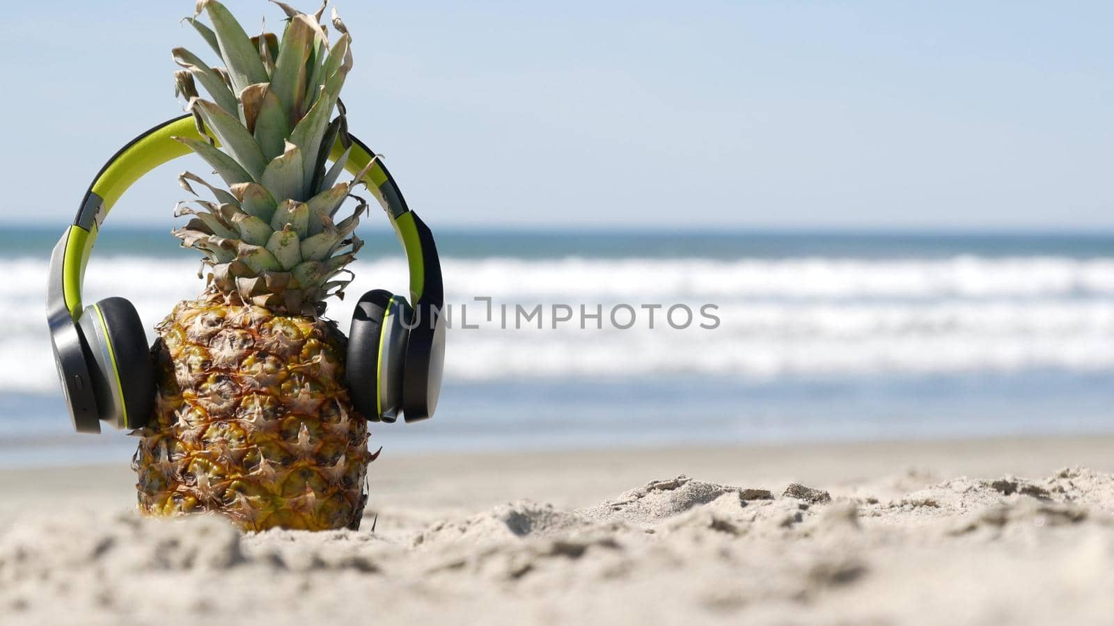 Funny pineapple in headphones, sandy ocean beach, blue sea water waves, California pacific coast, USA. Tropical summer exotic fruit enjoying vacations and music in paradise. Ananas sunbathing on shore