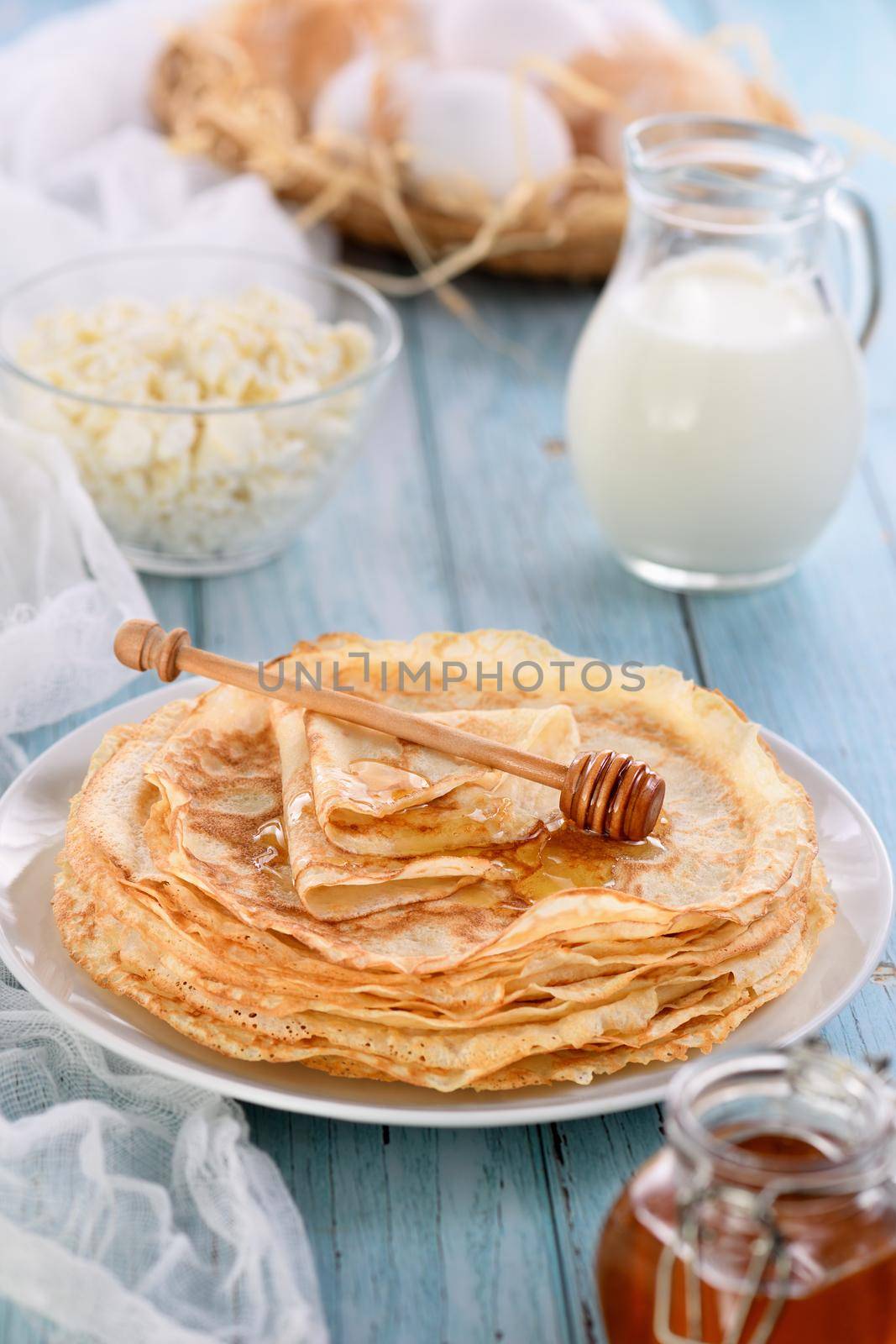 Homemade thin crepe (pancakes) with honey stacked in a stack, on a table with a pot of milk, a bowl of cottage cheese and eggs in a basket. Country style food. Traditional Slavonian, pagan holiday (Maslinitsa)