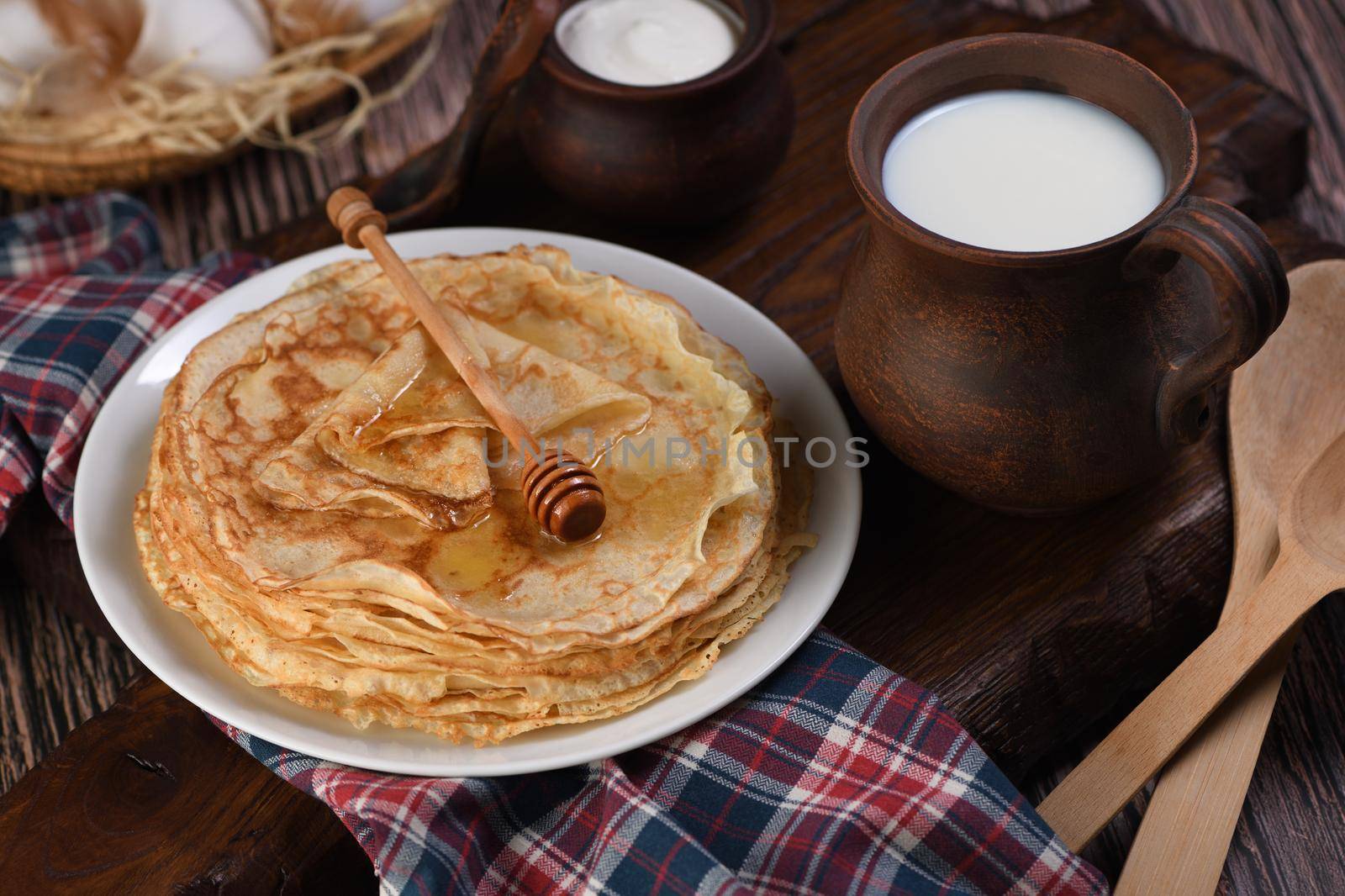 Homemade thin pancakes with honey stacked in a stack, on a wooden table with a mug of milk, a pot of sour cream and eggs in a basket. Traditional Slavonian, pagan holiday (Maslinitsa). Country style food.