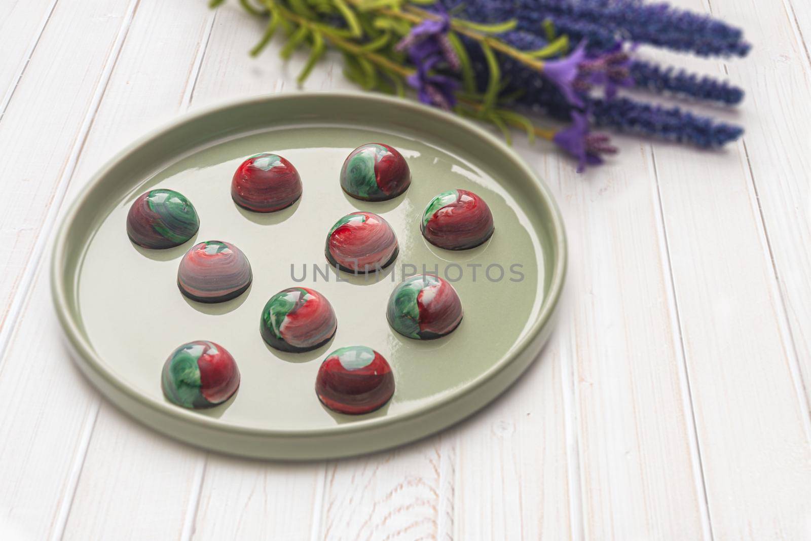 Collectible handmade tempered chocolate candies with a glossy painted body on a round plate with a blurred background and bokeh elements. Stock photography.