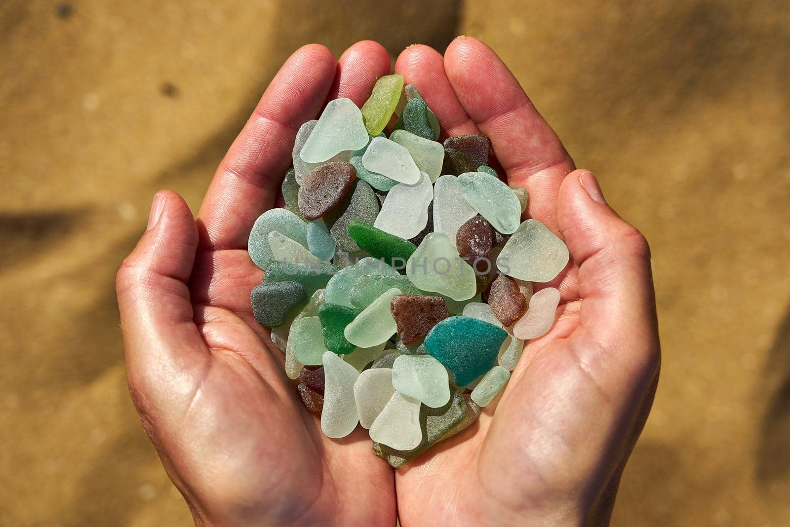 A person holds a collection of multi colored sea glass in their cupped hands with the sand in the background by ChrisWestPhoto