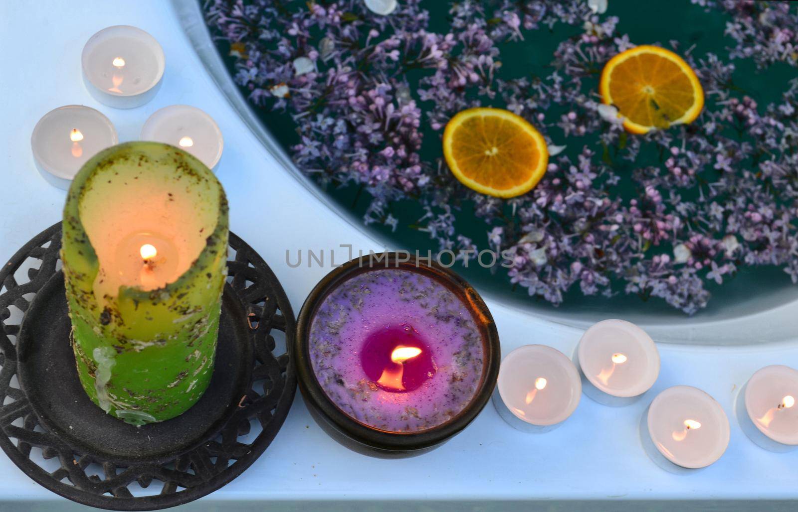 Colored candles on the edge of the bathtub full with blue water and lilacs flowers by hibrida13