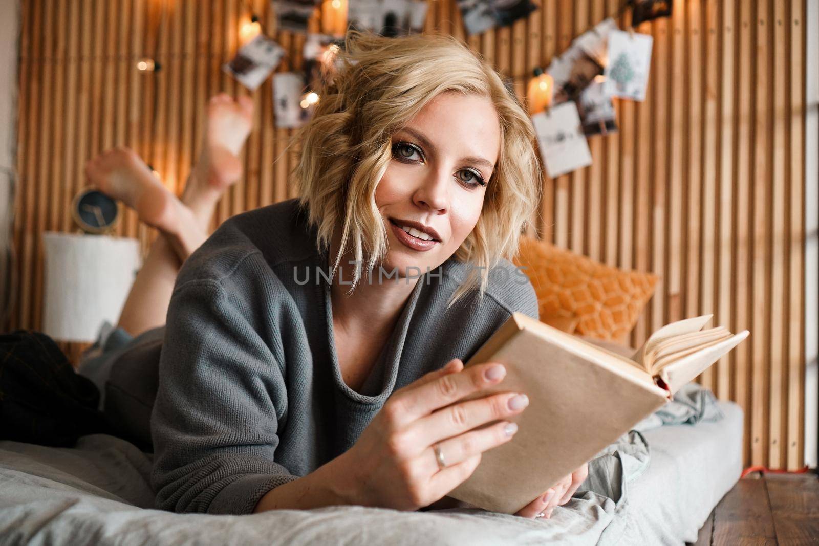 Happy Woman looking to the camera, relaxing and reading book on cozy bed - Wooden wall and photos with lights - blurred background