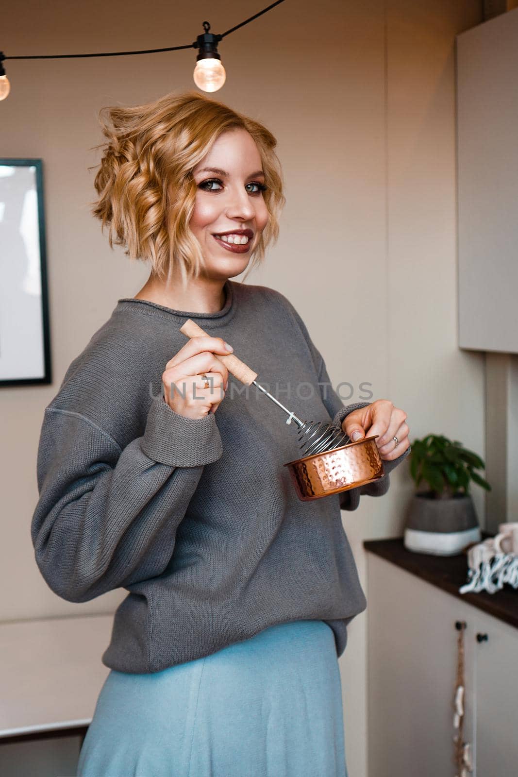 Smiling young woman in casual wear standing at the kitchen, holding ladle and whisk