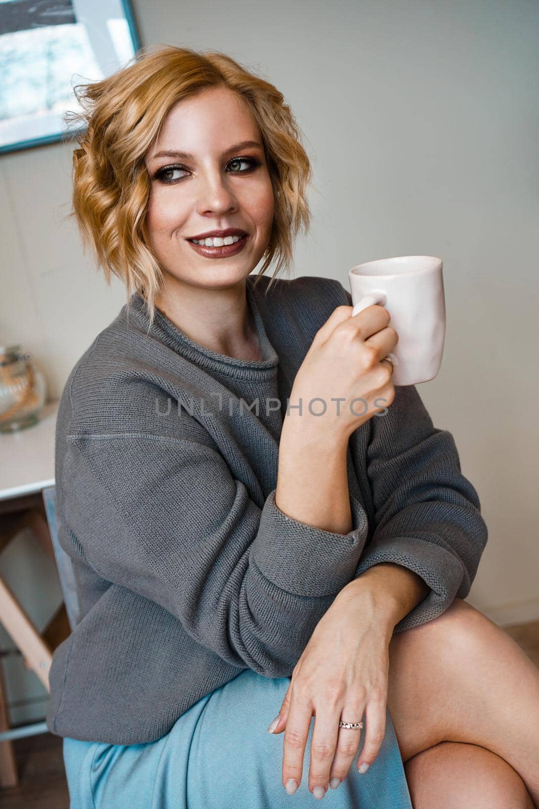 Smiling blonde drinking cappuccino, holding coffee cup. Vertical photo. Stay at home, cozy kitchen