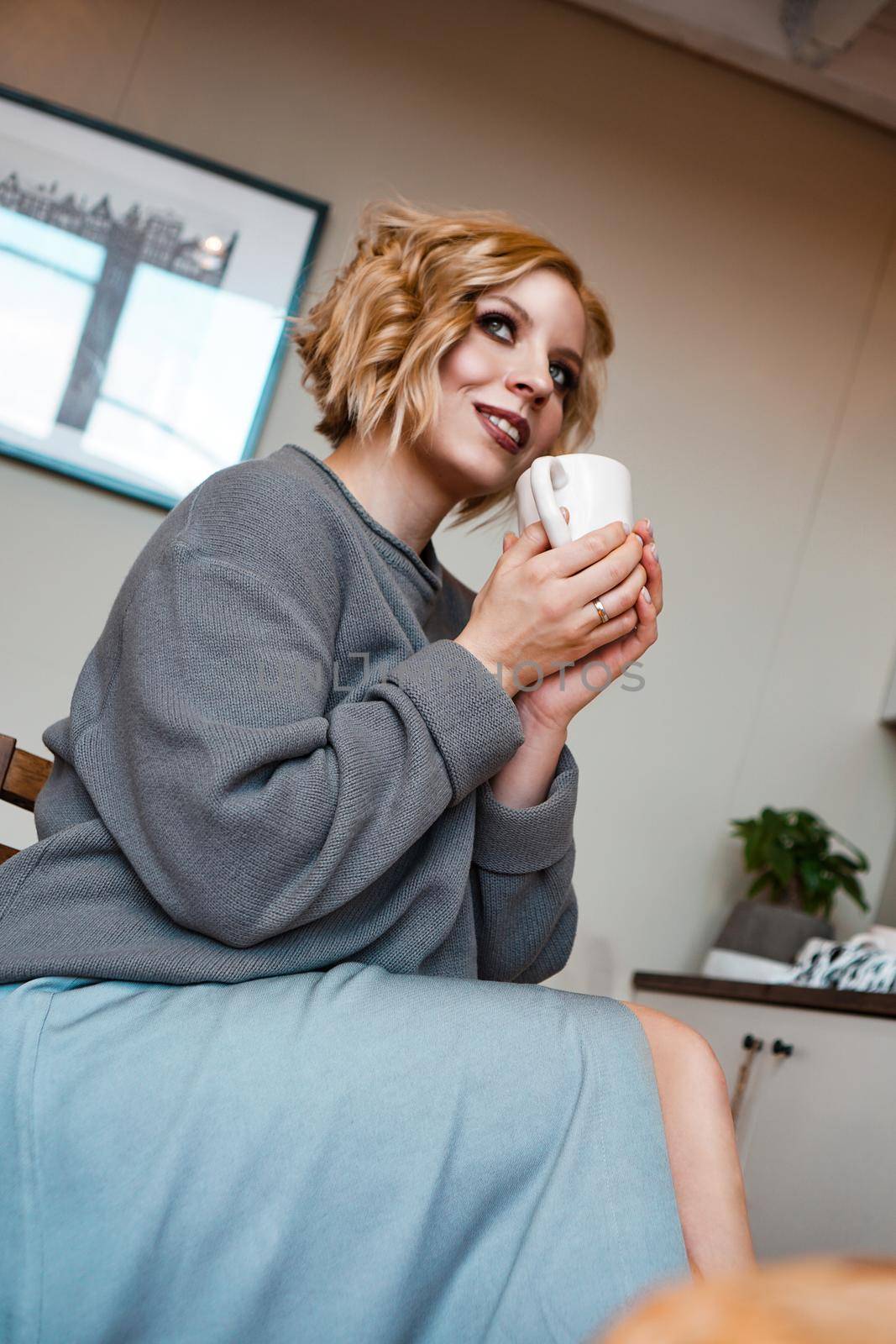 Smiling blonde drinking cappuccino, holding coffee cup. Vertical photo. Stay at home, cozy kitchen