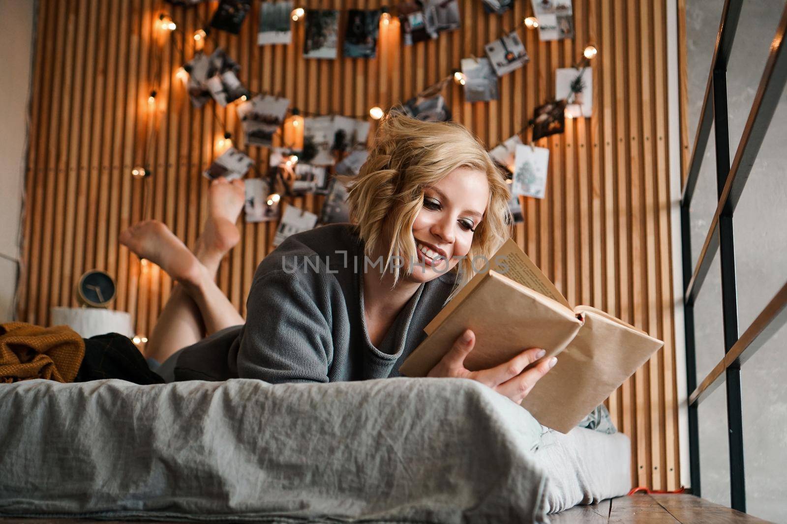 Happy Woman relaxing and reading book on cozy bed - Wooden wall and photos with lights - blurred background