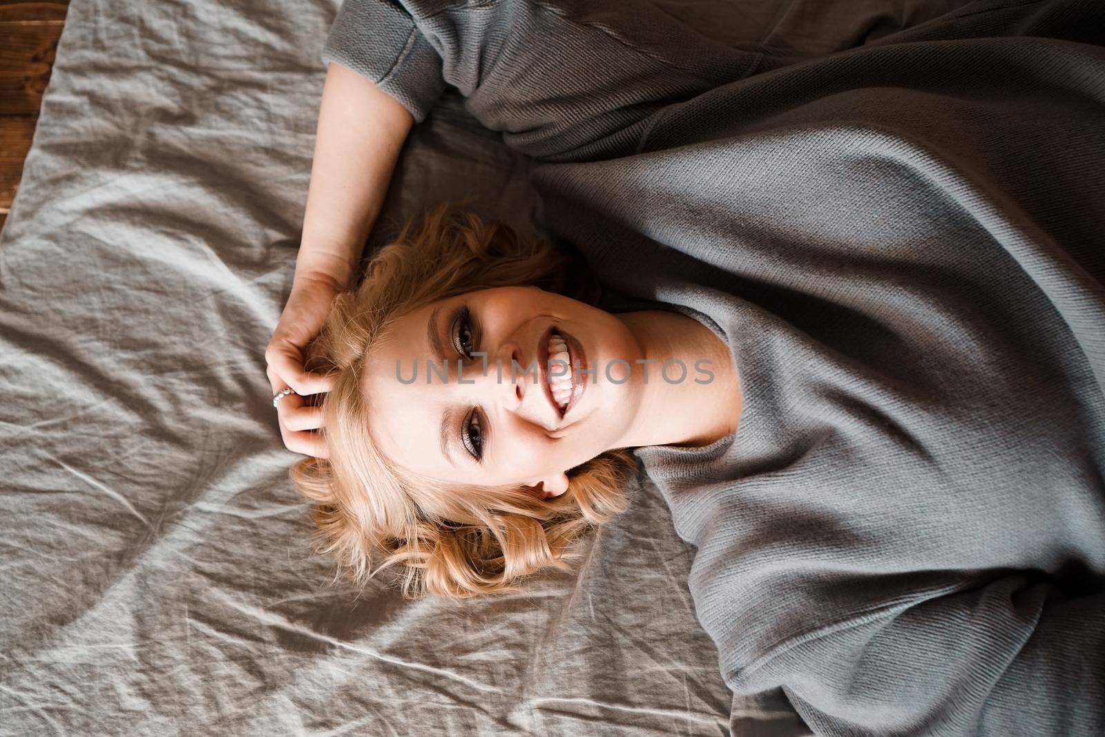 Beautiful young woman in gray sweater laughing lying in bed in her cozy bedroom. Cute girl relaxing at home. Happy morning