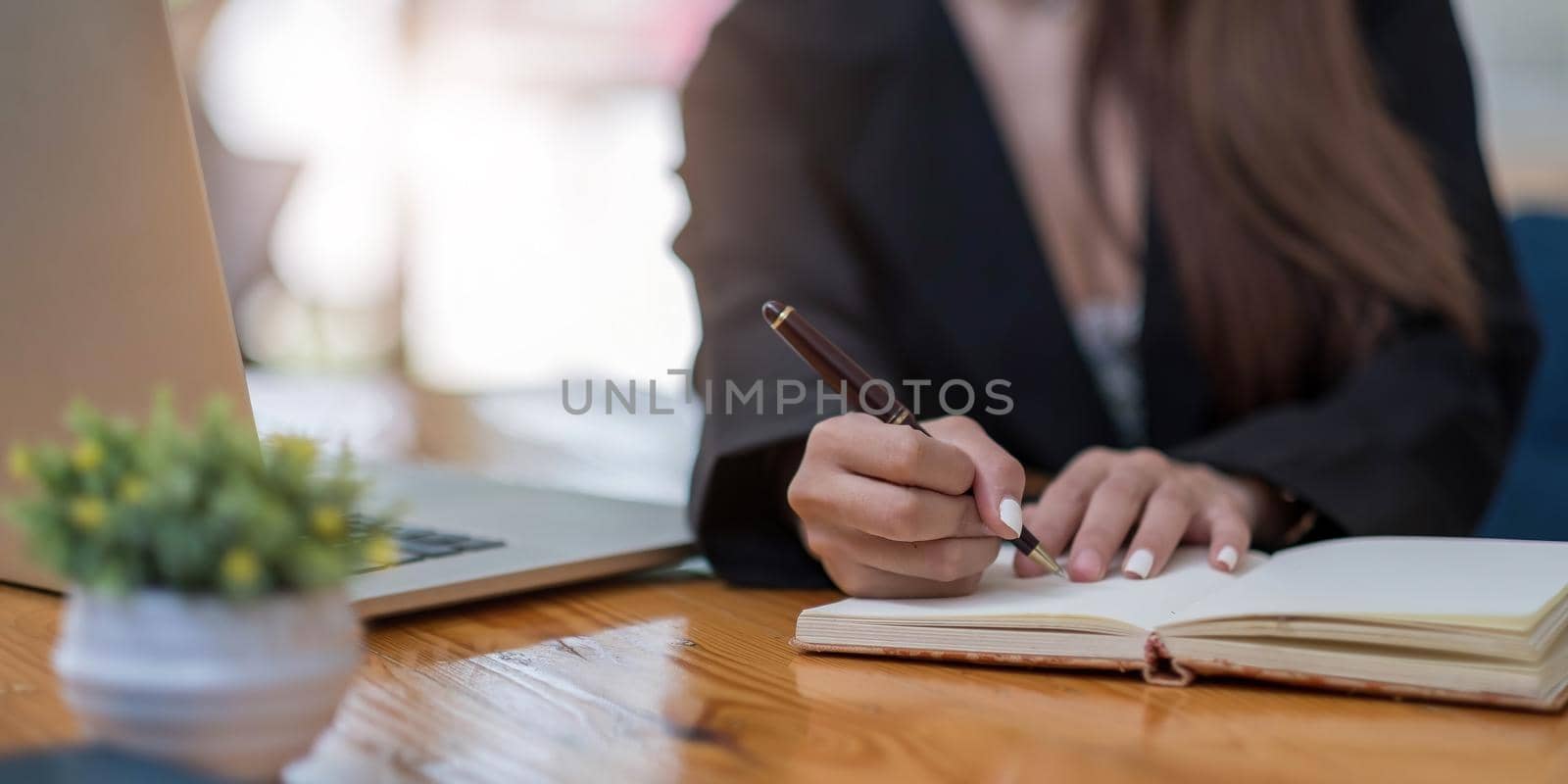 Close up woman's hands with laptop computer, notebook and pen taking notes in business office by wichayada