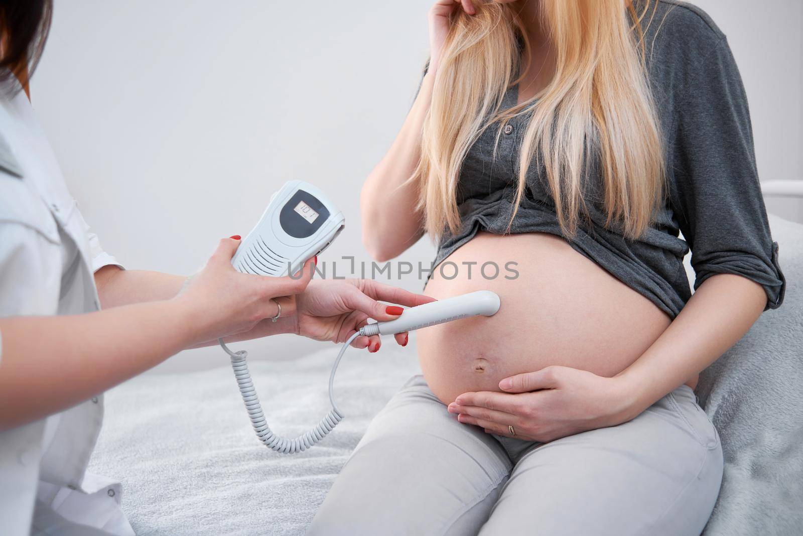 Crop of female doctor in white diagnosing pregnant woman, touching stomach , checking and listening heart beat of baby with special tool. Patient at procedure of diagnostic in hospital.