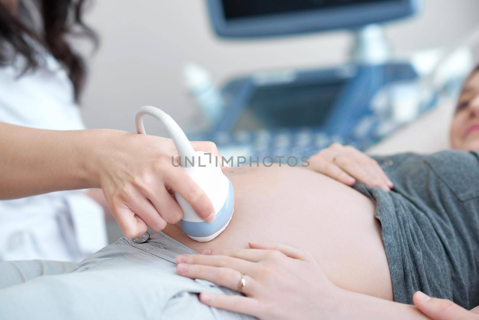 Close up of incognito professional doctor using ultrasound equipment, screening of pregnant woman. Doctor in white uniform touching stomach by tool of young female, who lying on bed at cabinet.