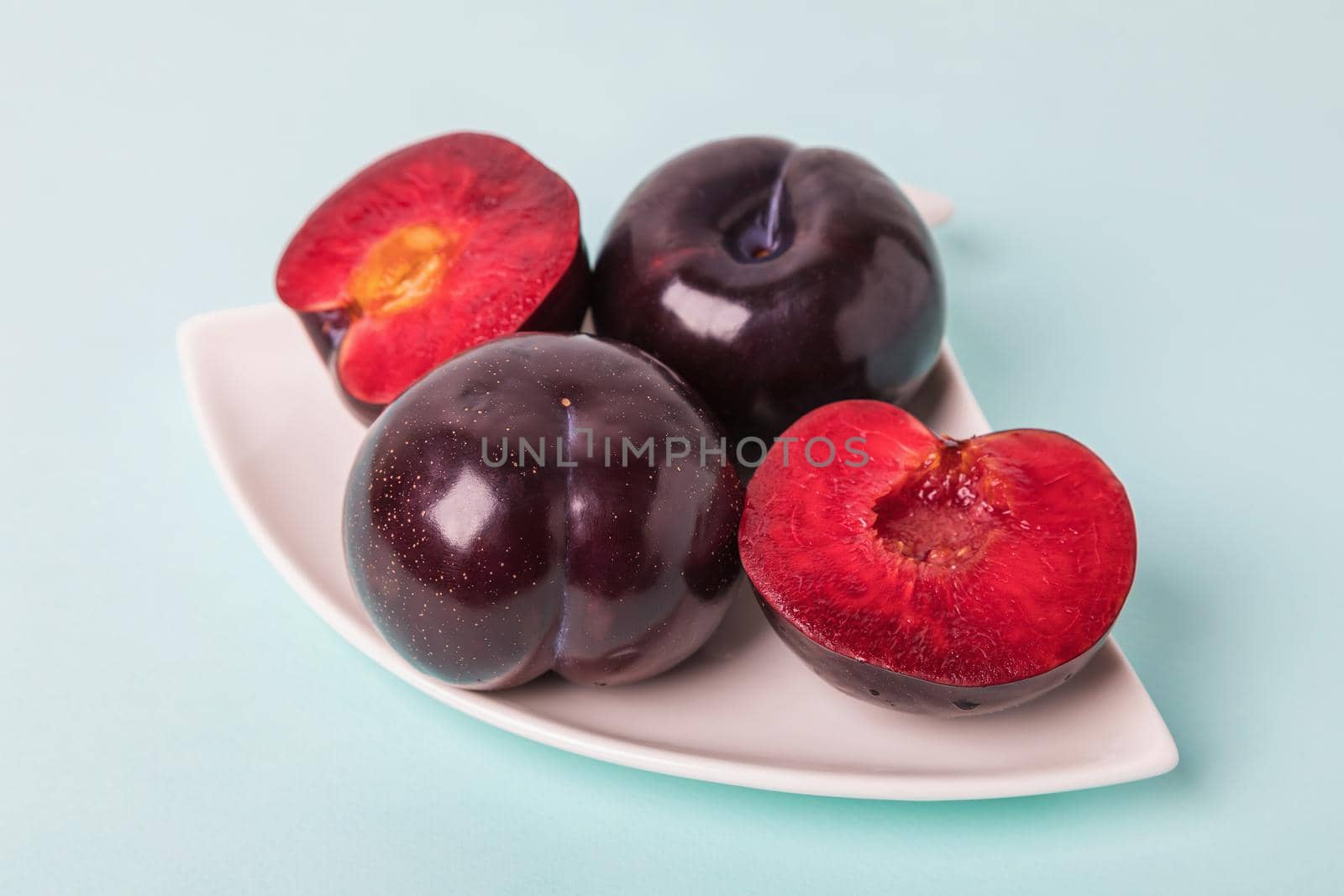 On a white saucer is a large plum, cut in half, and next to it is a whole plum. The pulp of red berries. Close-up.
