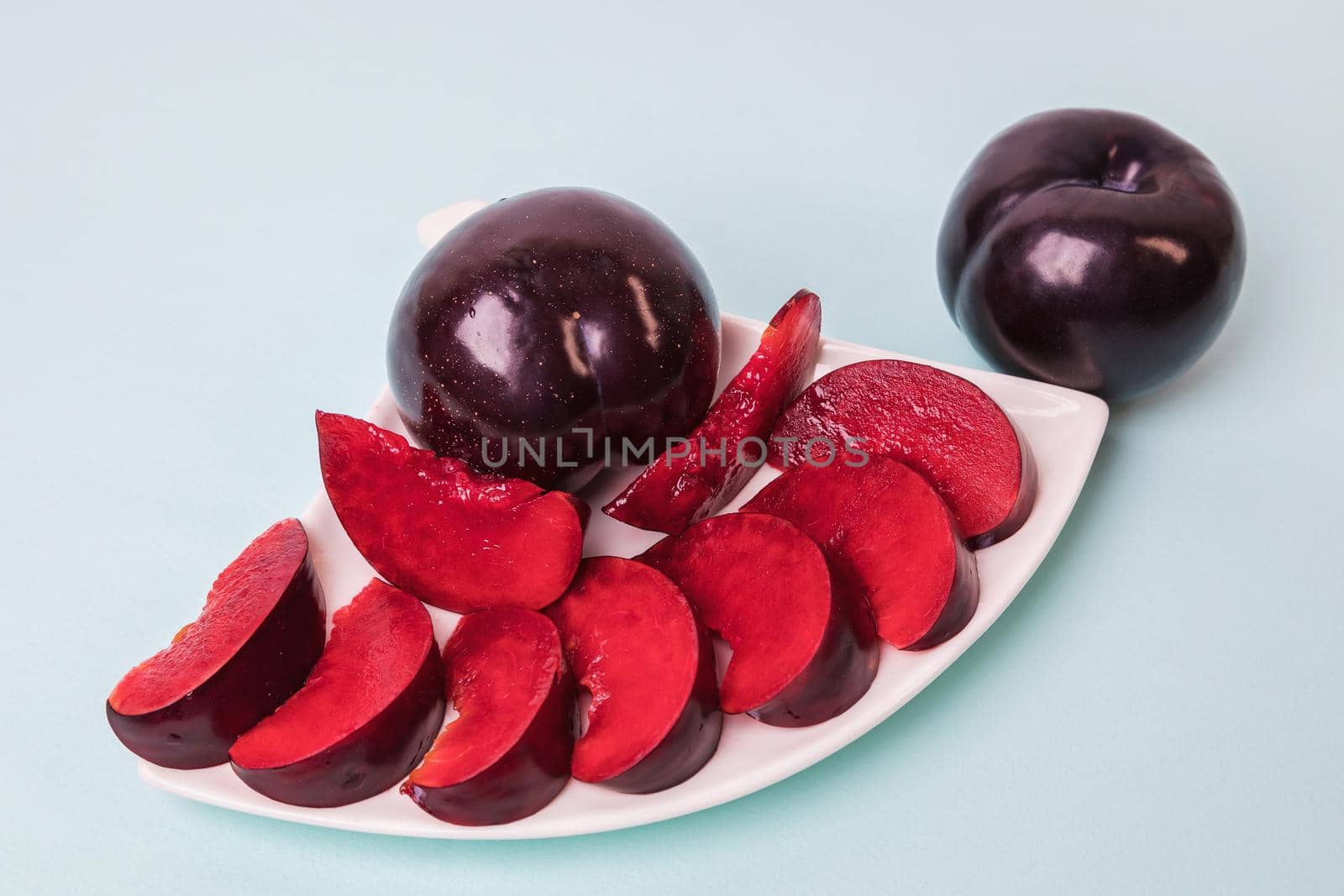 On a white saucer there is a large plum cut into wedges, next to it there are two more plums. The pulp of red berries. Close-up.