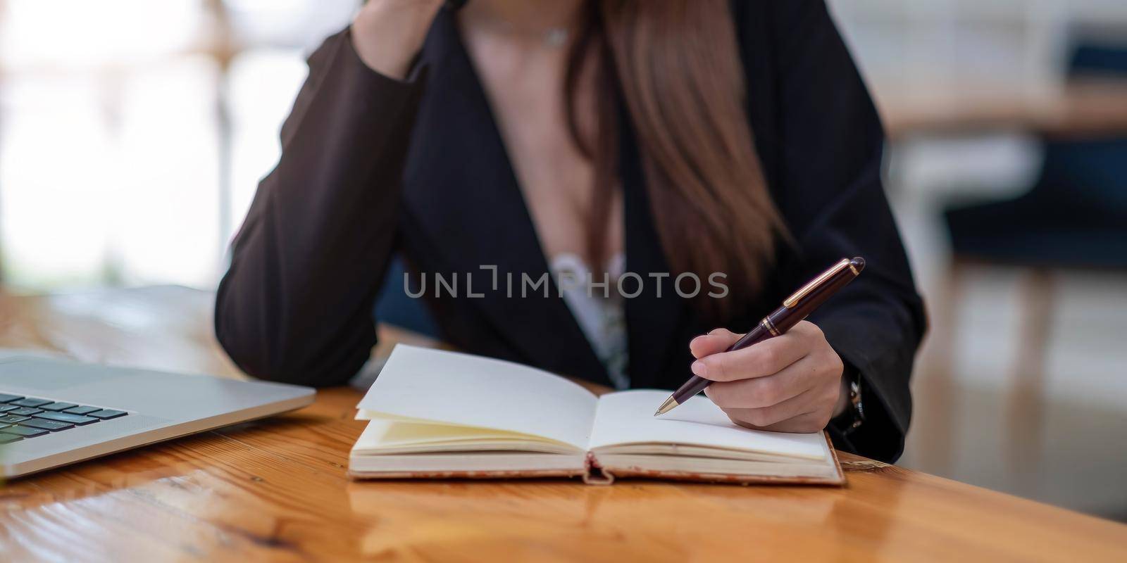 Close up woman's hands with laptop computer, notebook and pen taking notes in business office by wichayada
