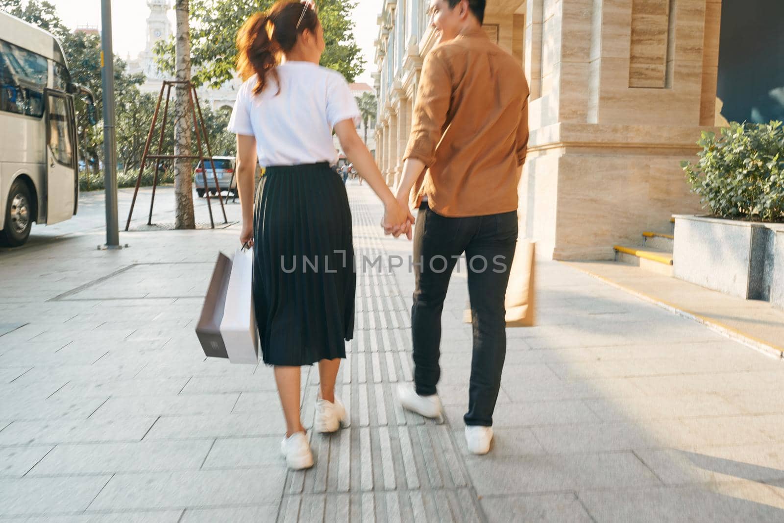 Lower body section of a young tourist couple walking by store windows and holding paper shopping bags in a destination city.