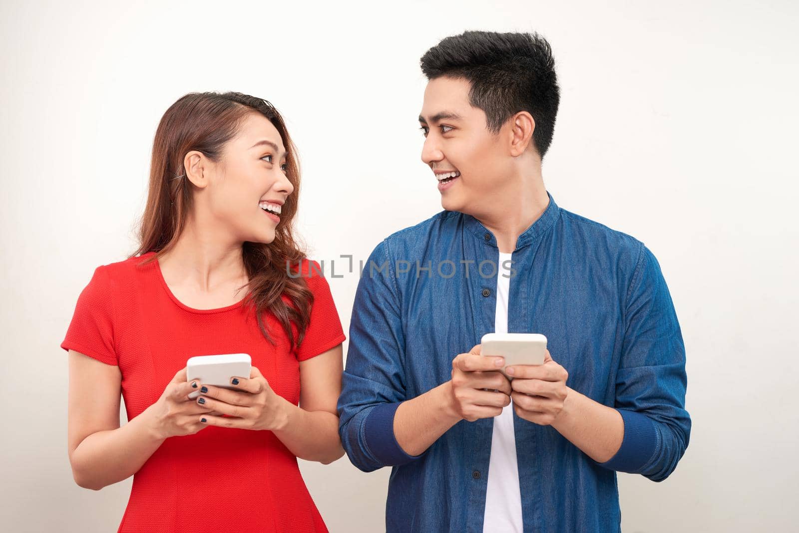 Beautiful smiling modern couple in casual wear with phones in hands on a white wall background by makidotvn