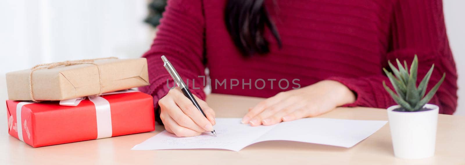 Closeup hand of young asian woman writing postcard in Christmas day at home, eve and celebrate, female writing message on greeting card with giving gift box in holiday, congratulation and celebration.
