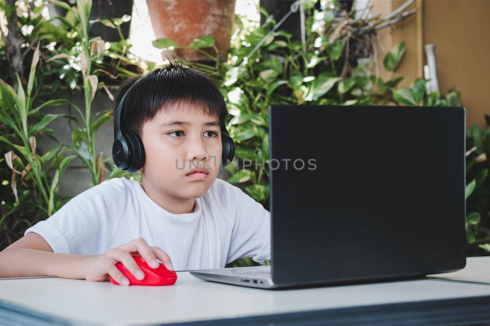 Asian boy wears wireless headphones and uses laptop computer to study online. by wattanaphob
