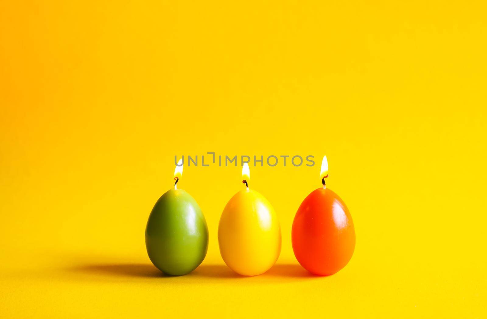 Traditional Easter decor. Group of bright burning paraffin candles in the shape of colorful eggs on soft warm yellow background.