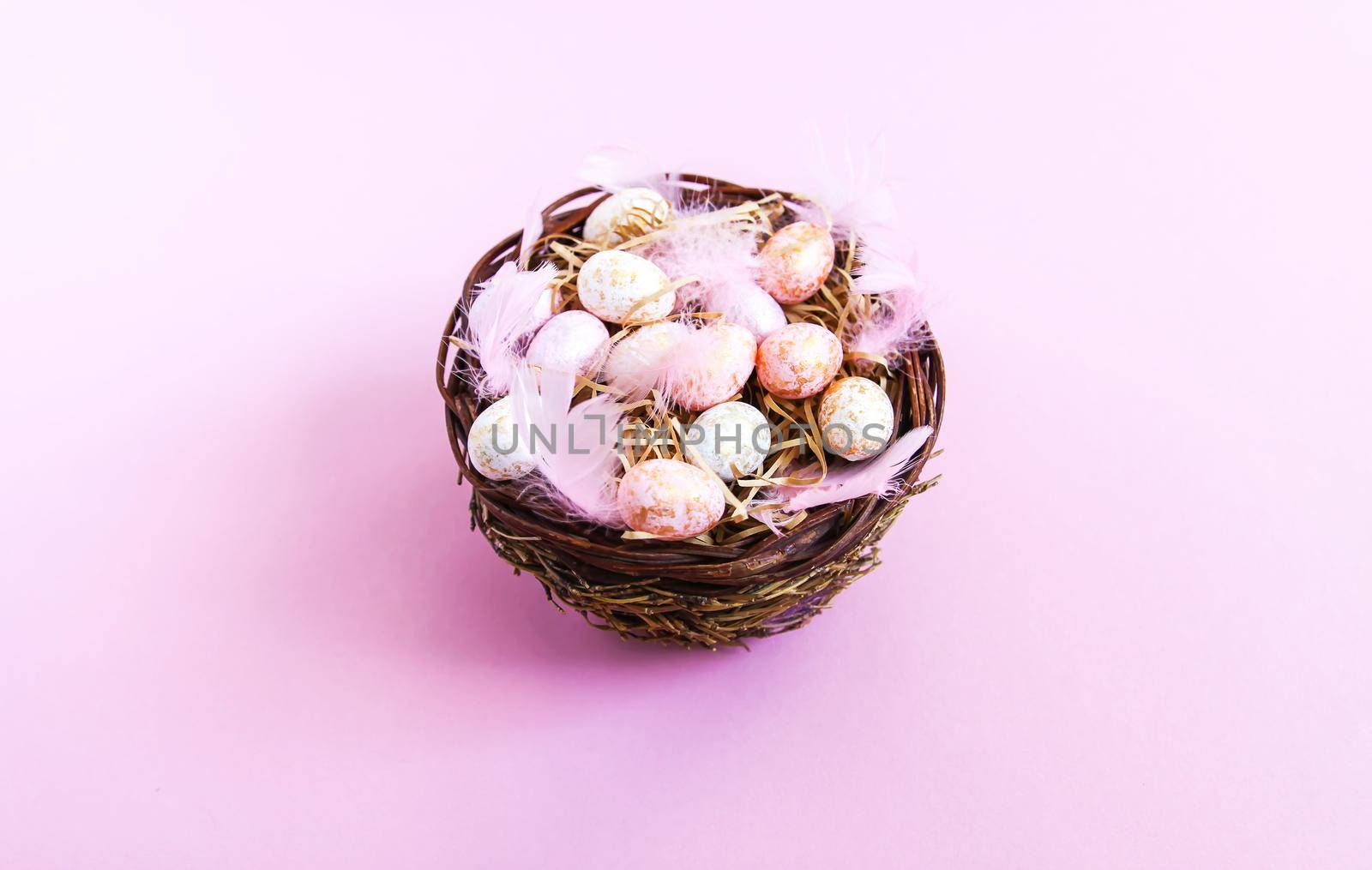 Easter composition with traditional decor. Small decorative colorful eggs and soft feathers in a wicker basket on pink light background.