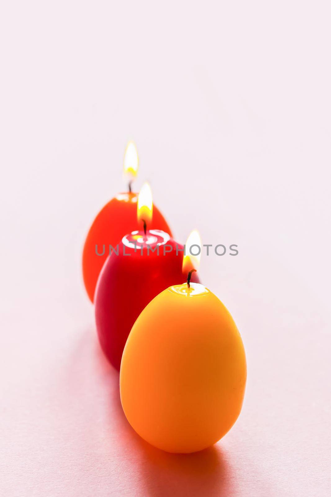 Traditional Easter decor. Group of bright burning paraffin candles in the shape of colorful eggs on soft pink background.