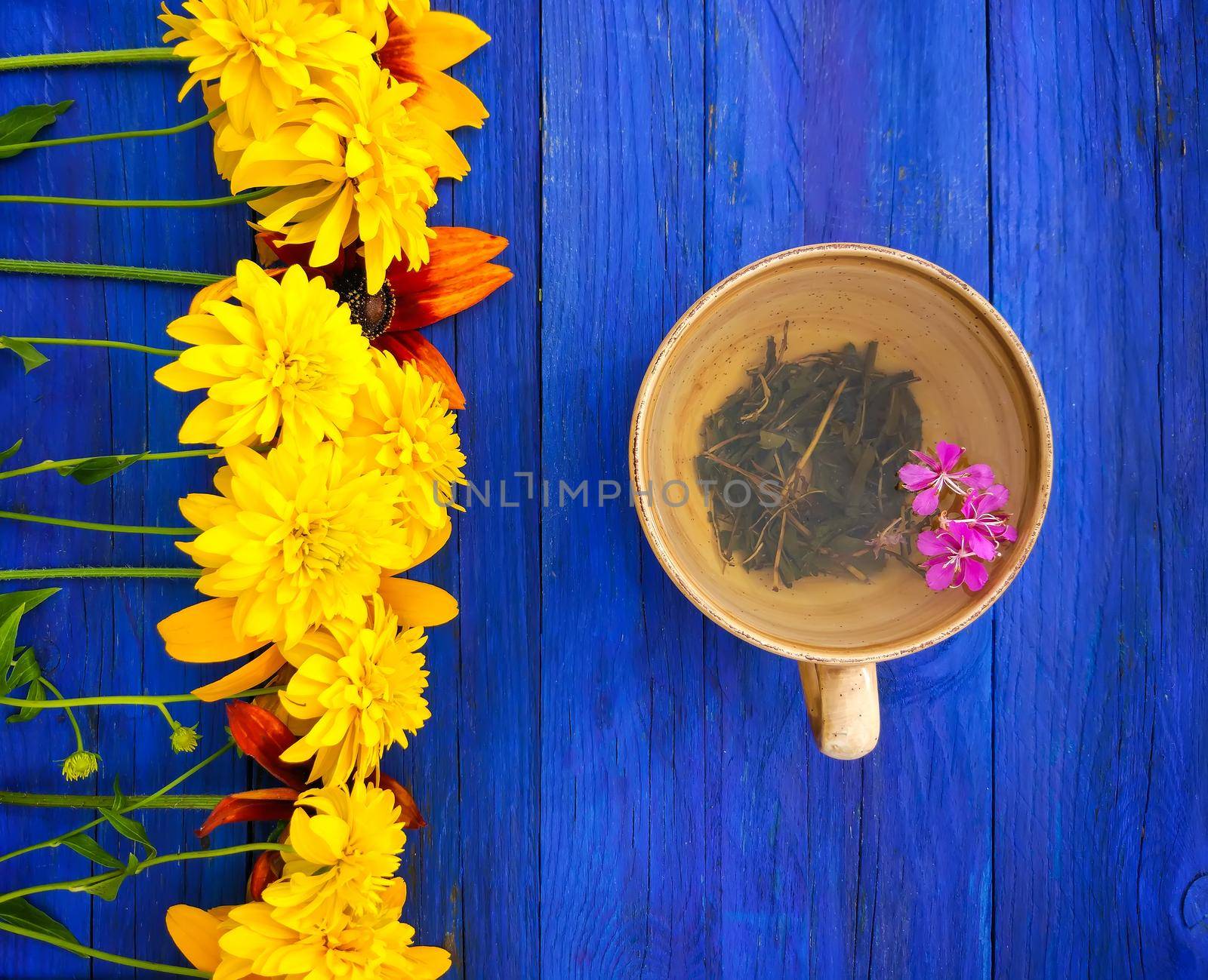 Natural herbal tea with purple fresh flowers and leaves of medical fireweed plant n ceramic cup on blue wooden boards outdoors.