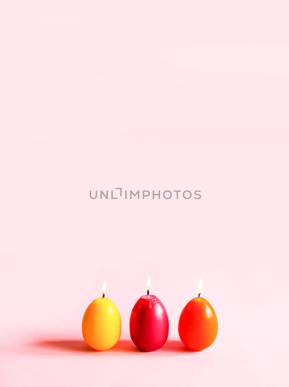 Traditional Easter decor. Group of bright burning paraffin candles in the shape of colorful eggs on soft pink background.
