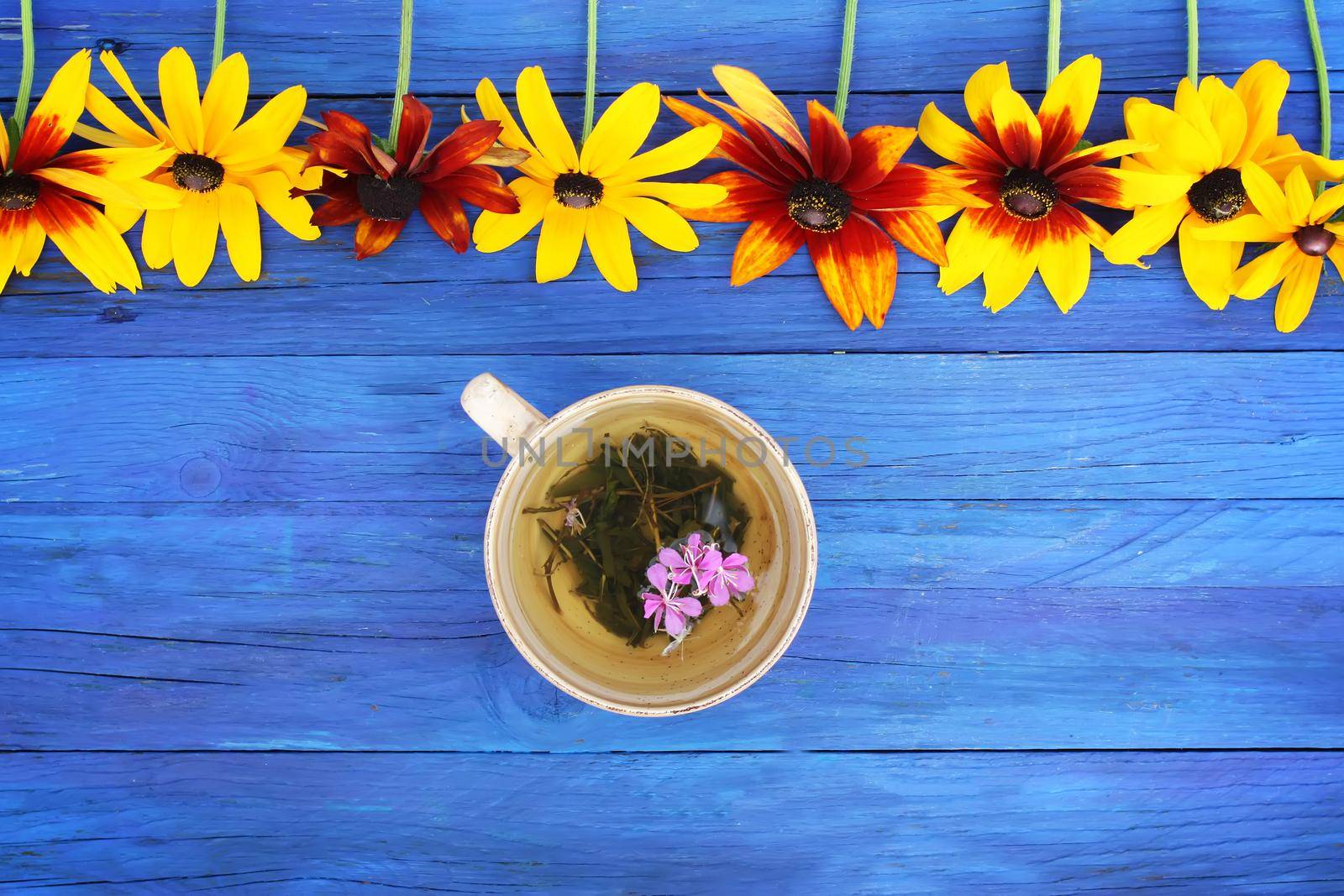 Natural herbal tea with purple fresh flowers and leaves of medical fireweed plant n ceramic cup on blue wooden boards outdoors.