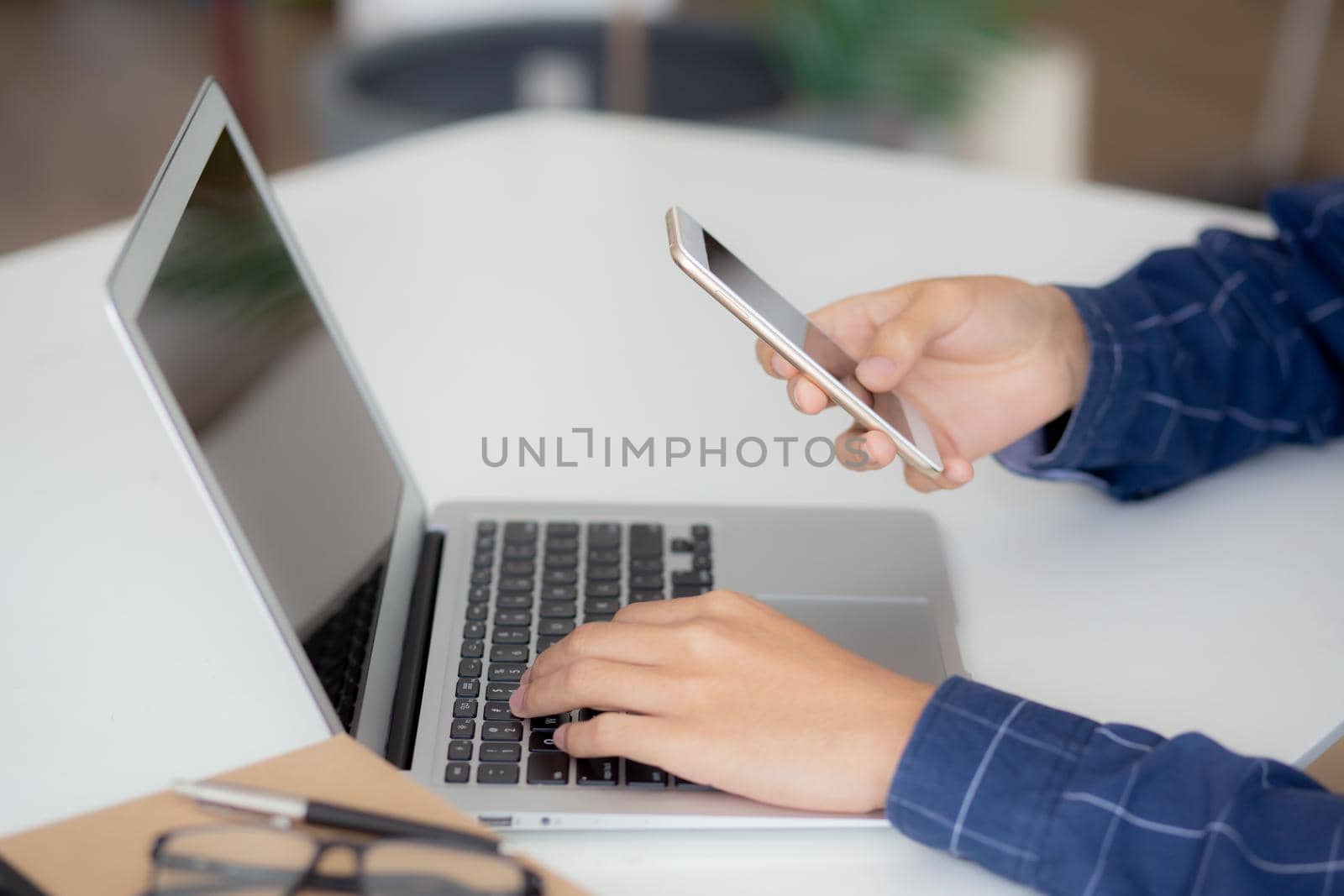 Closeup hand of young man working laptop computer and reading smartphone on internet online on desk at home, freelance male using phone with social media, business and communication concept.