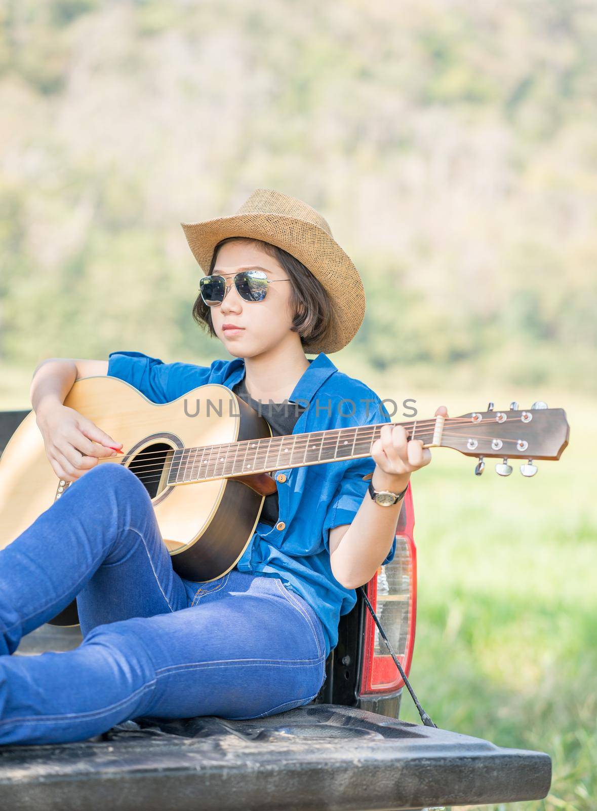 Young asian women short hair wear hat and sunglasses playing guitar ,sit on pickup truck in countryside Thailand