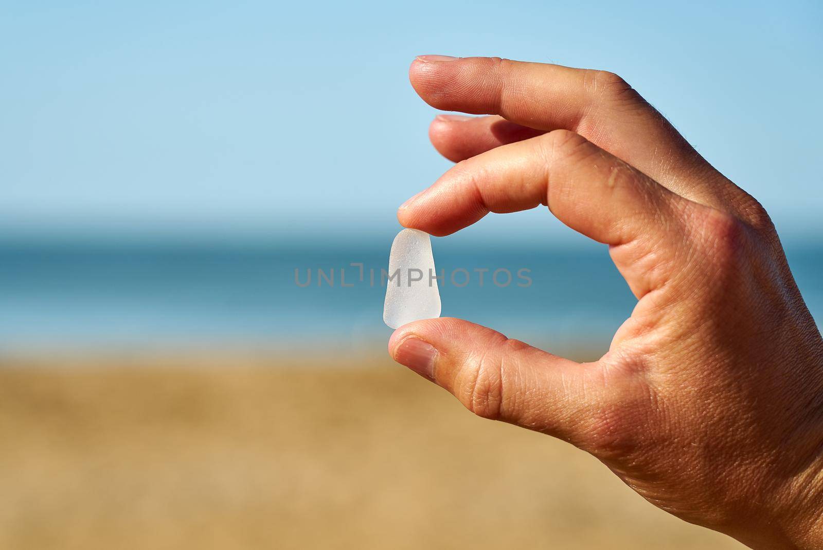 White frosted sea glass is held up with the sand, sea and sky in the background