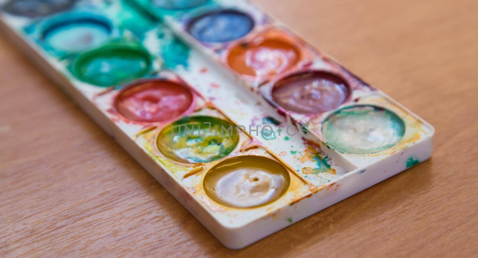 On the table is a used set of multicolored watercolor paints for painting. Top view of half-empty jars of watercolors. Flat layout on the table.