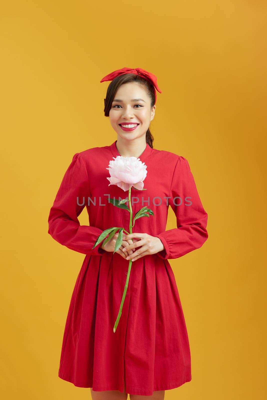 Portrait of adorable young woman standing with charming smile, holding peony flower in hands and smells it.