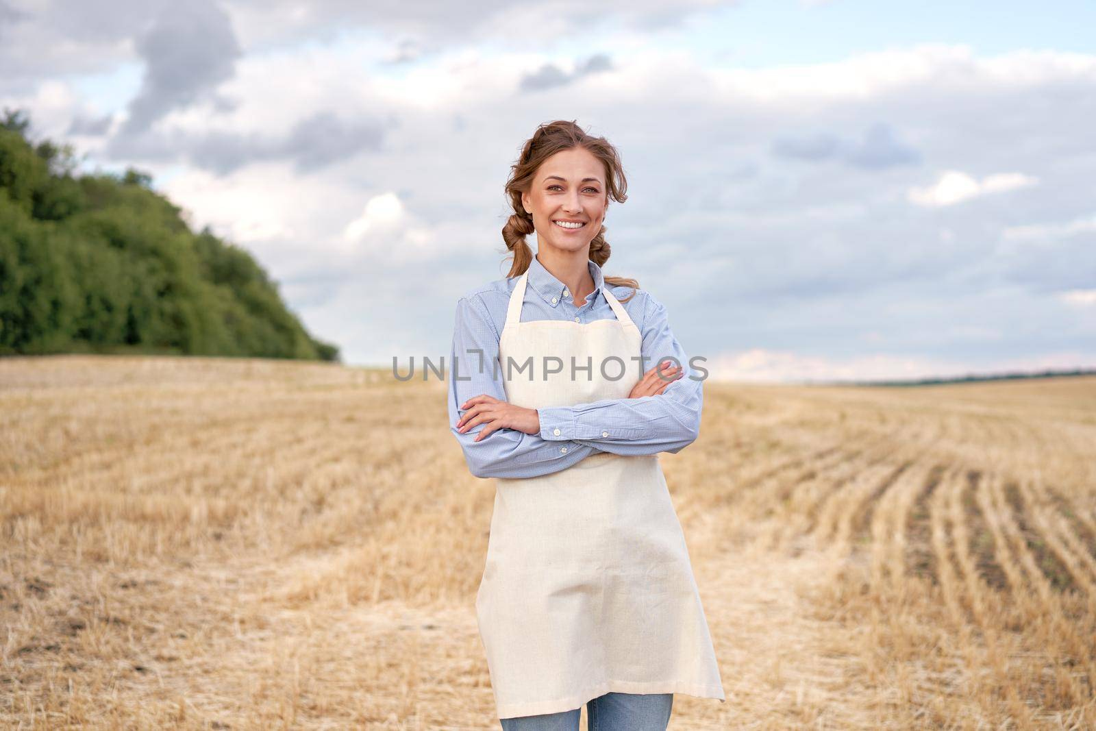 Woman farmer apron standing farmland smiling Female agronomist specialist farming agribusiness Happy positive caucasian worker agricultural field Pretty girl arms crossed cloudy sky background