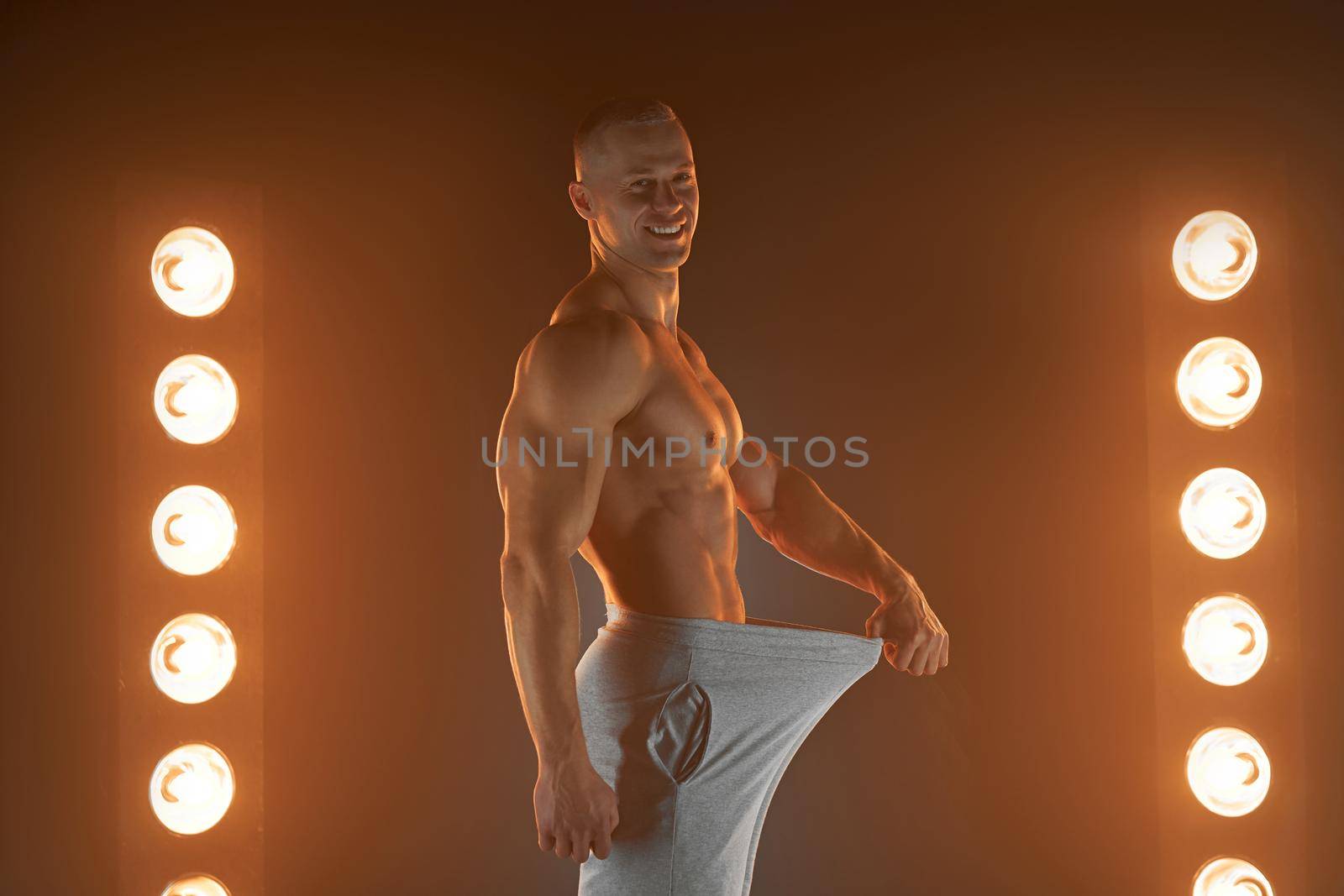 Great male size. Young muscular guy pulling back his pants and showing thumb up gesture, being proud and satisfied with his genitals, lamps illumination on background Weight loss concept Diet