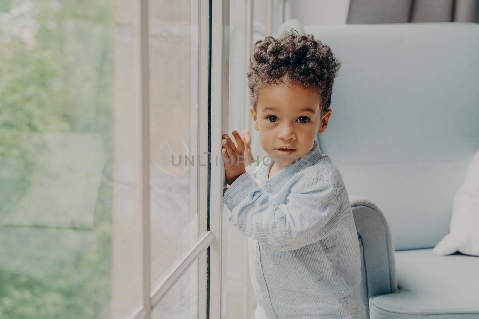 Portrait of adorable afro american curly haired baby boy in light blue colored shirt leaning on window glass in morning sunlight in living room at home while waiting for parents. Childhood concept