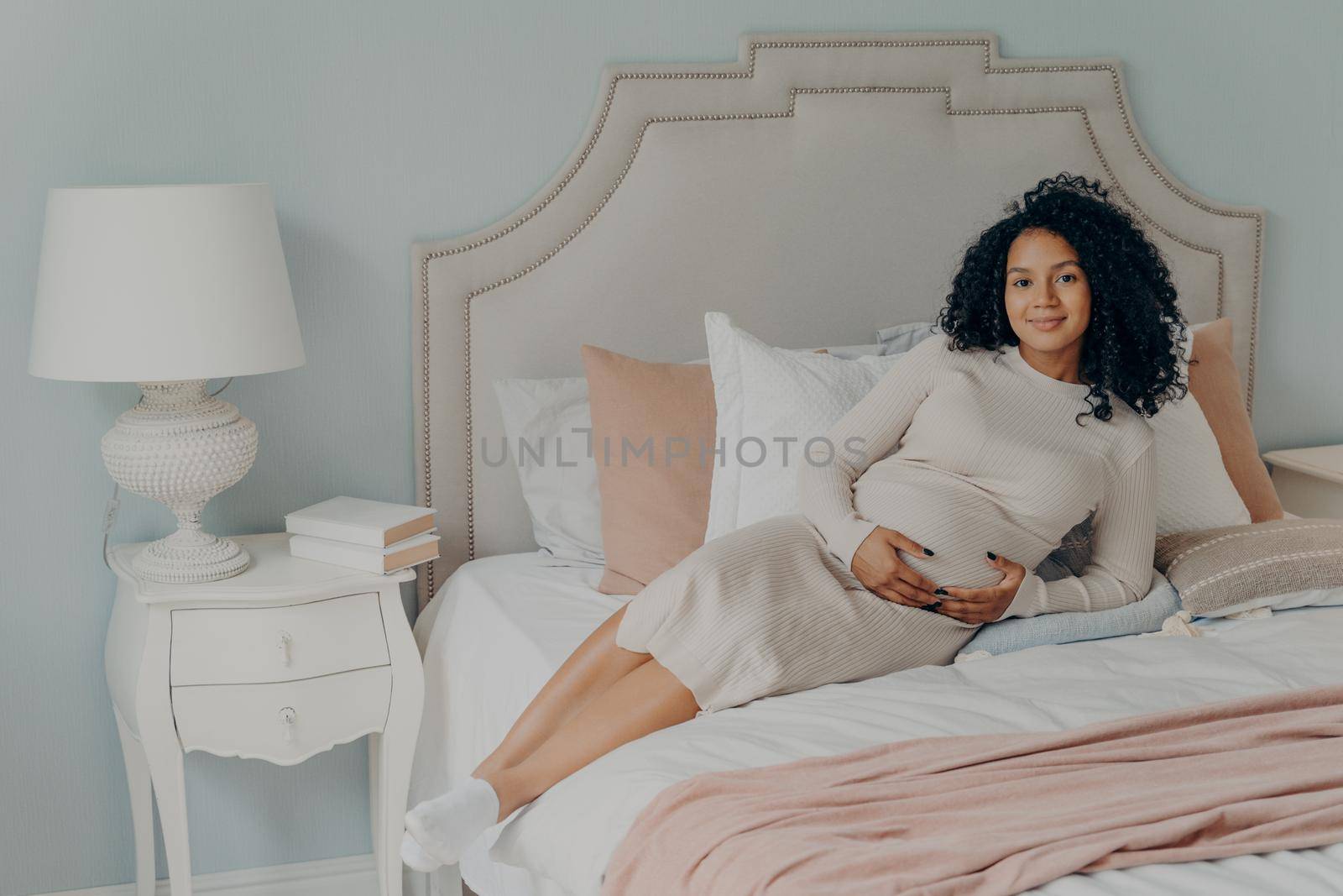 Happy lovely pregnant female relaxing on bed at home and touching her belly by vkstock