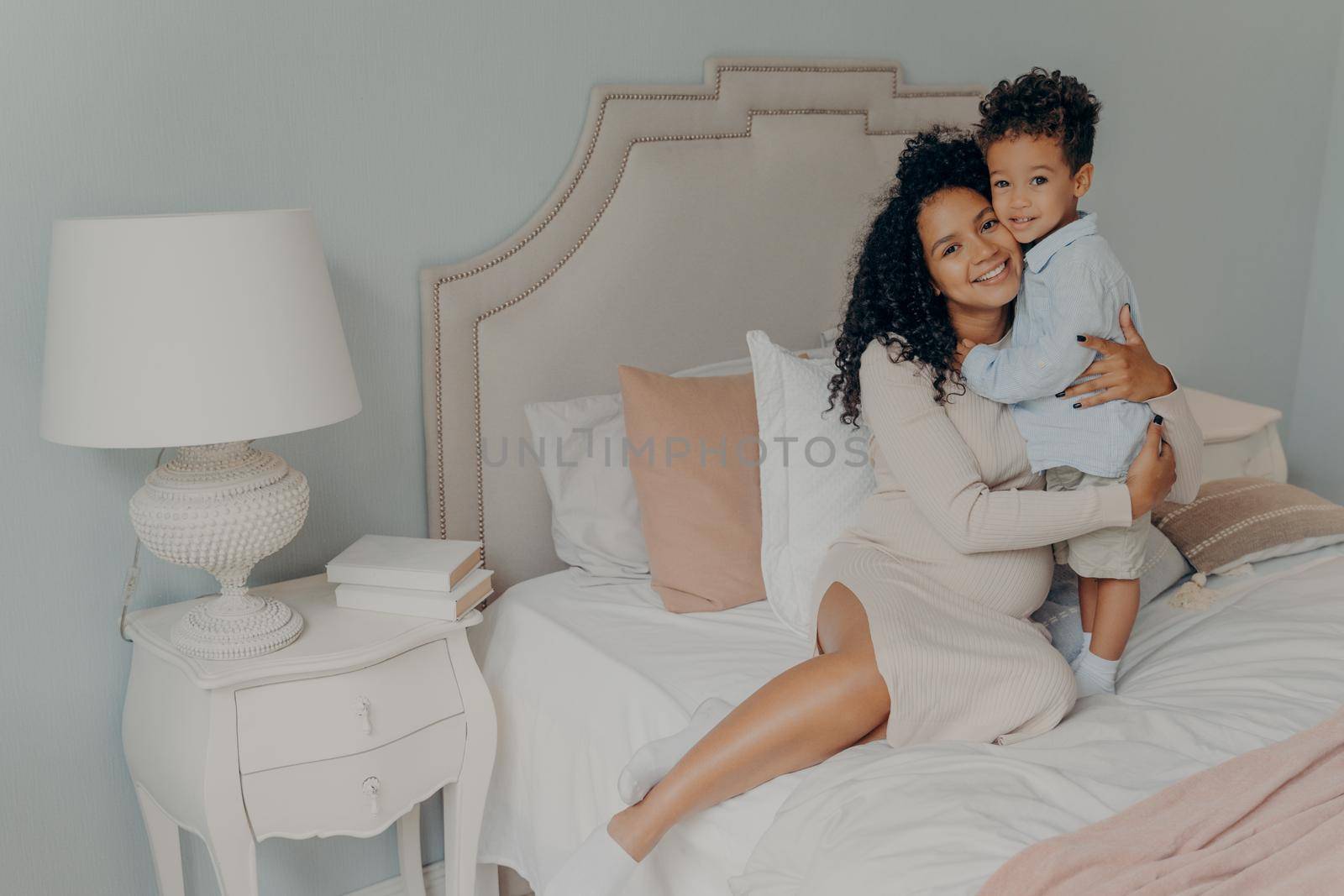 Happy pregnant mixed race lady expecting another child and enjoying pregnancy while sitting on spacious bed and hugging little cute boy son while spending time together at home. Motherhood concept
