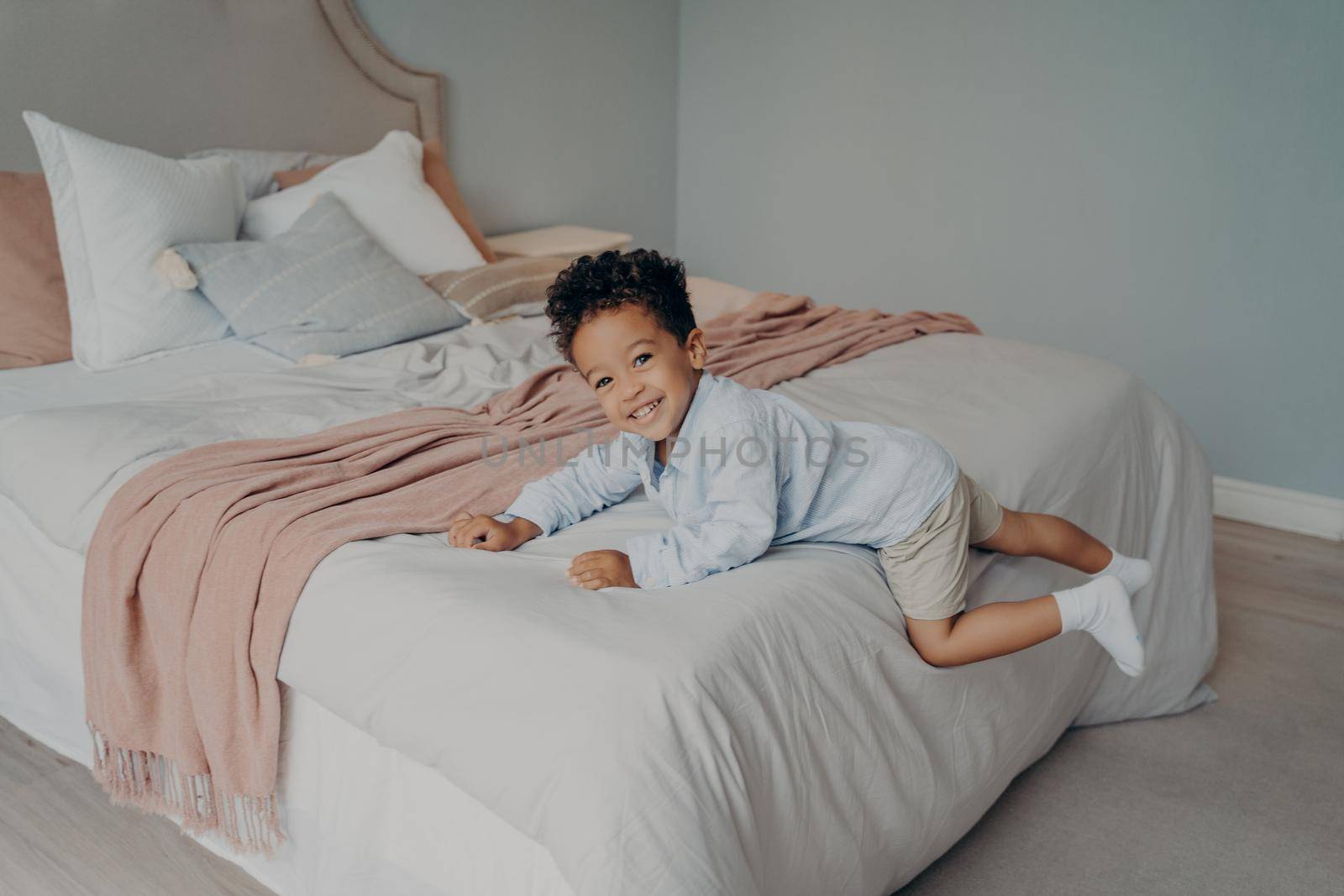 Joyous little mixed race boy kid laying and resting on his big comfortable bed with pillows, feeling happy and smiling while playing in bedroom. Children spending leisure time at home